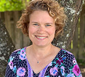 Lori Therrien in a purple shirt with flowers stands outside in front of a tree