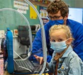 elementary student looks at the camera while a teacher looks over her shoulder_166x150px