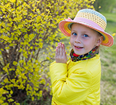 child in yellow coat and pastel hat stands outside next to a blooming bush