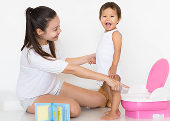 toddler girl stands next to a small pink and white potty with a woman nearby