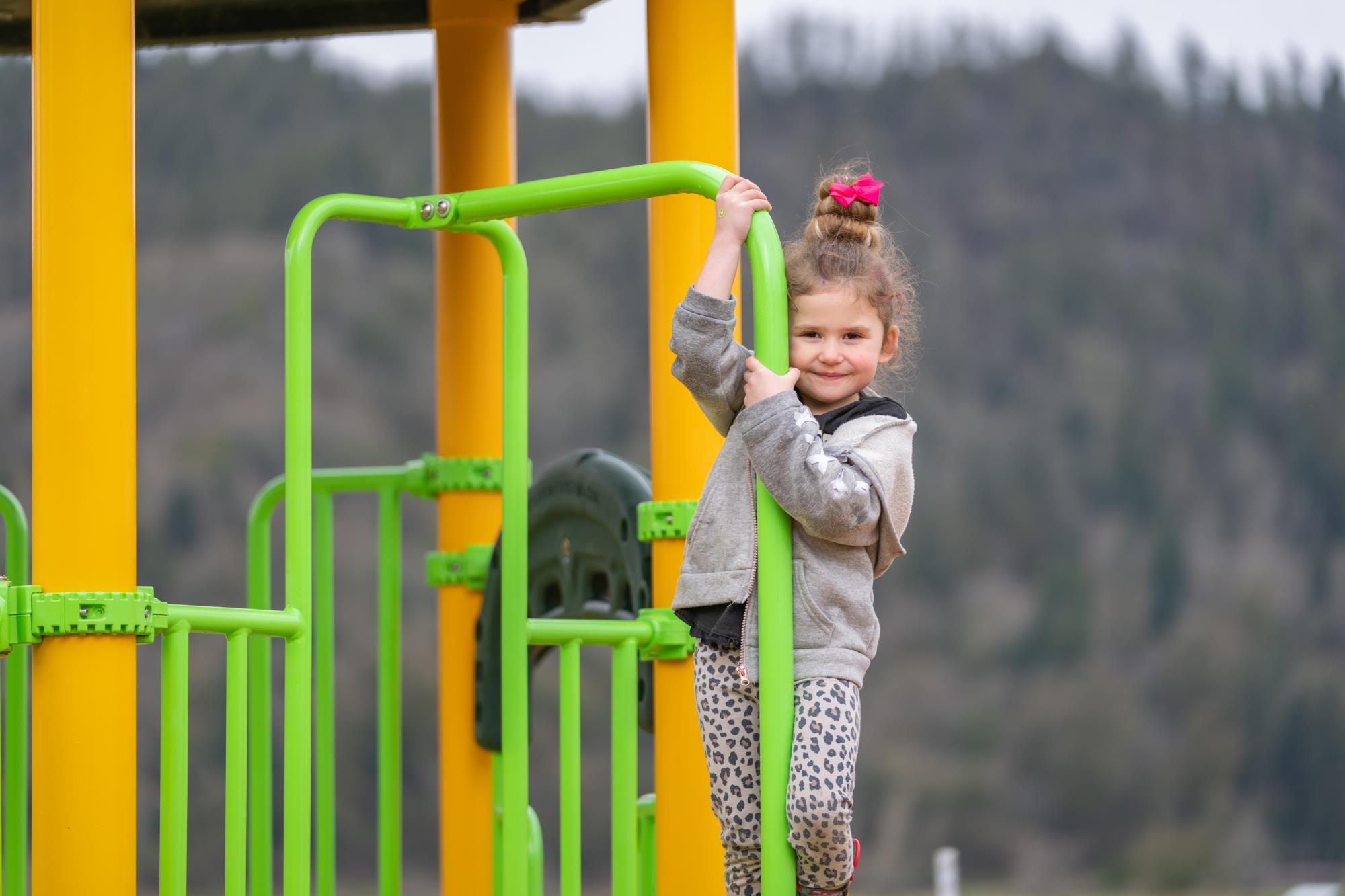 A preschool student holds onto a lime green bar on the playground with forest in the background