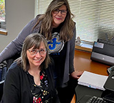 two women pose for a photo at a desk