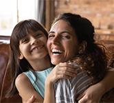 a girl and her mom sit on the couch at home and smile at the camera