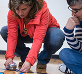 a teacher in a red jacket holds a round robot while another teacher looks on