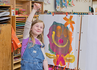 a preschooler holds her hand up in the air after she finishes a painting on an easel