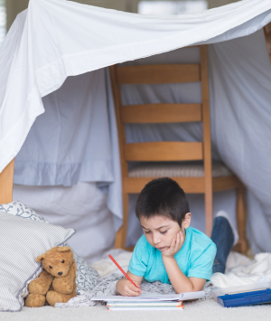 Boy writing with a pencil lays under a blanket fort next to a teddy bear