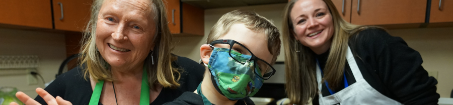 Kid in colorful face mask with glasses poses with two adults in aprons in front of kitchen cabinets in a cooking class