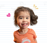 Toddler in an orange shirt with brown hair smiles for the camera