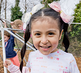 a preschooler with pink bows in her hair stands on the playground