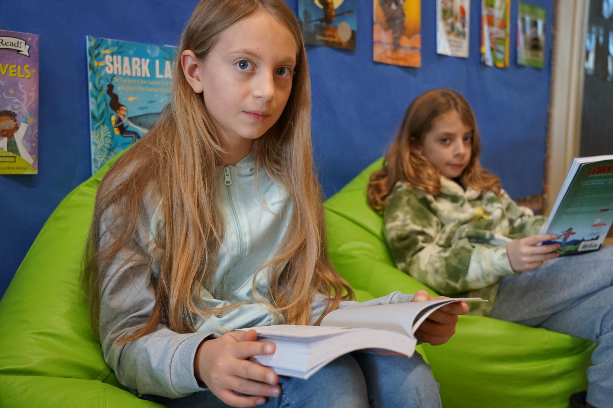 Paige reads a book in the library while Elijah reads nearby