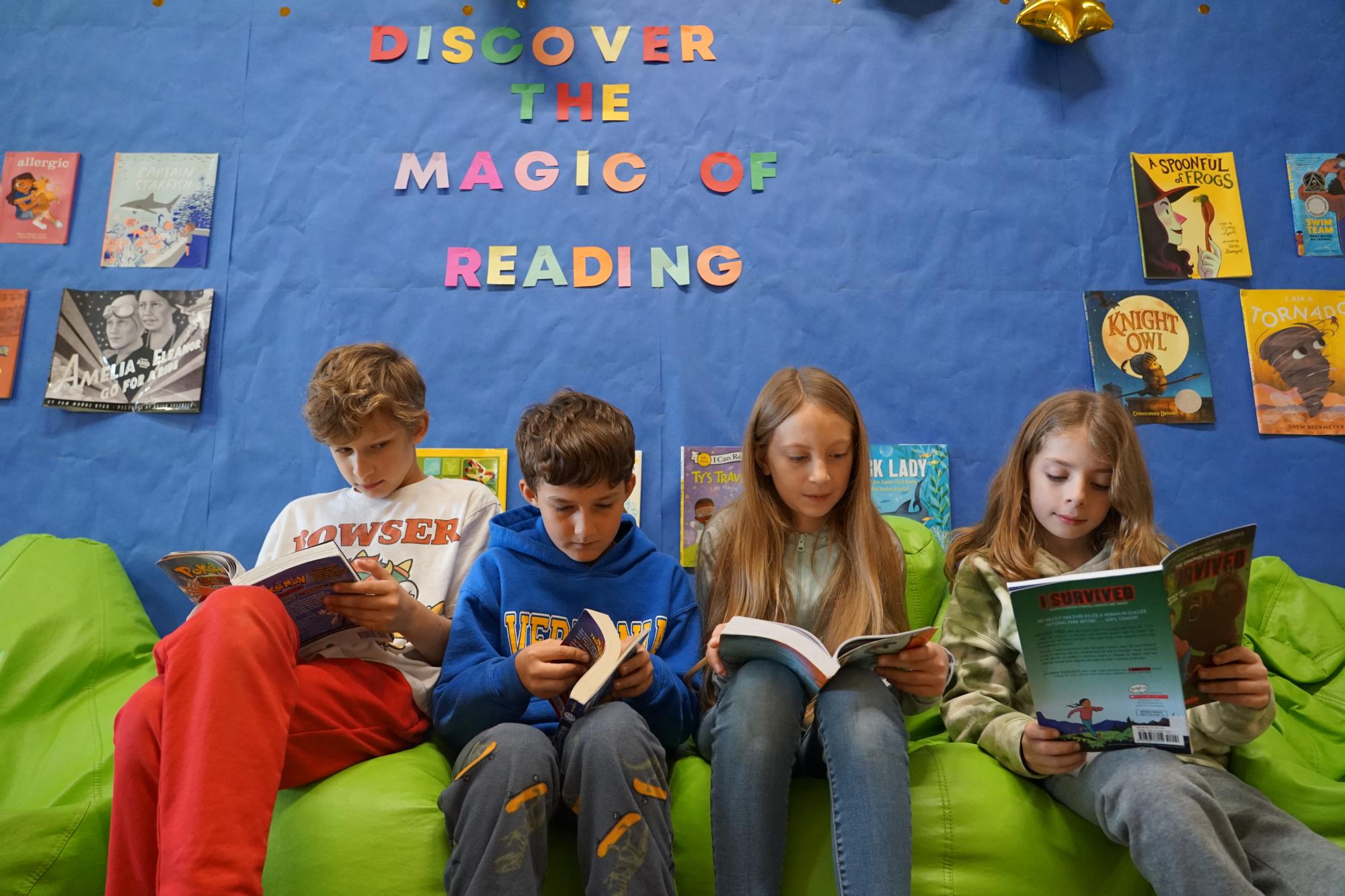 Four students read books on green bean bags in the library with book titles behind them and text that says discover the magic of reading