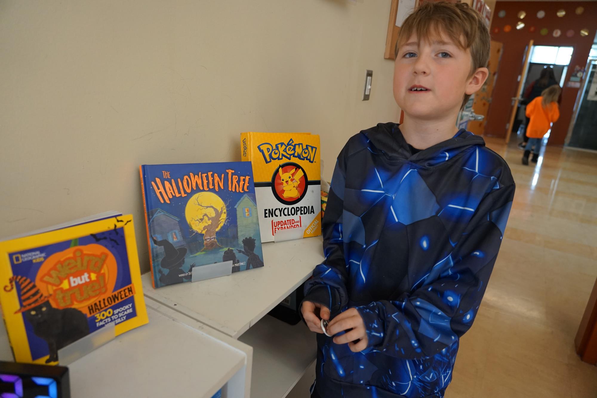 Cole wears a black and dark blue hoodie and stands next to a book shelf in his classroom