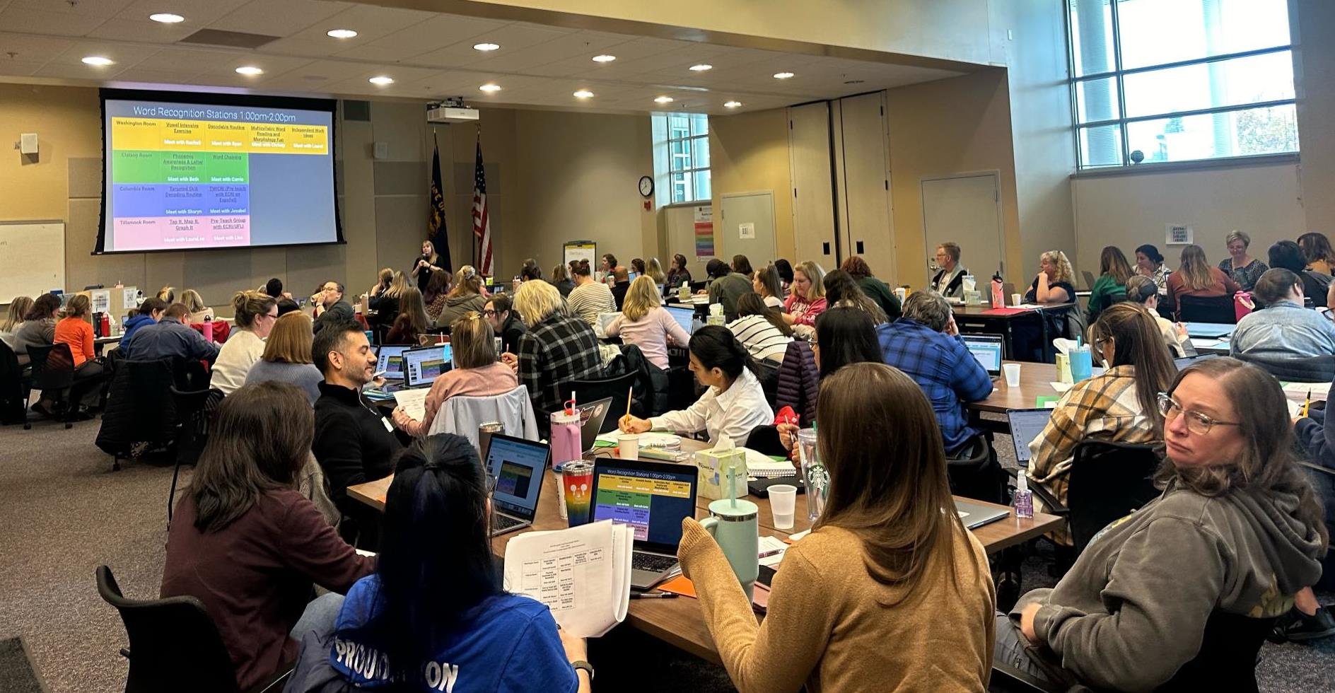 Teachers sit at rectangular tables in a large conference room